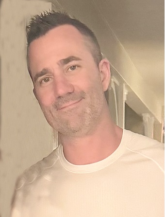 Smiling man with short dark hair and light stubble, wearing a white crew-neck shirt, looking at the camera indoors.