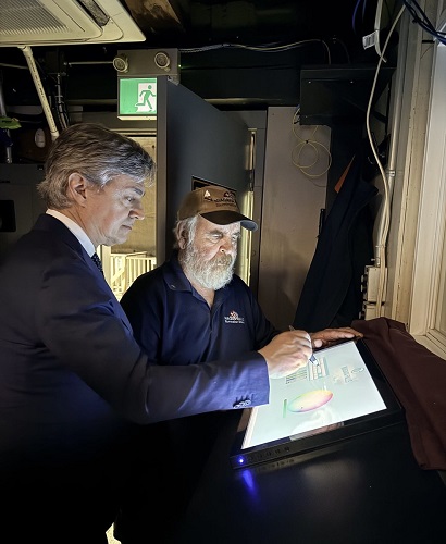 Two men, one in a dark suit and the other in a cap, stand at a touchscreen kiosk, touching the screen in a dim backstage area near an exit sign.