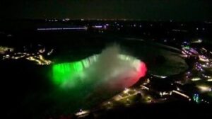 Aerial night view of Niagara Falls illuminated in green, white, and red lights with surrounding city lights on the shore.