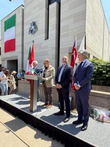 Public ceremony outside a modern government building, with the Italian flag and other banners; a man speaks from a podium while three officials stand nearby as onlookers watch.