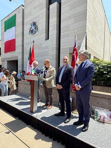 Public ceremony outside a modern government building, with the Italian flag and other banners; a man speaks from a podium while three officials stand nearby as onlookers watch.