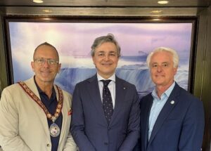 Three men in formal attire posing for a photo; left wears a ceremonial chain, middle in a dark suit, right in a blue blazer, with a waterfall painting behind.