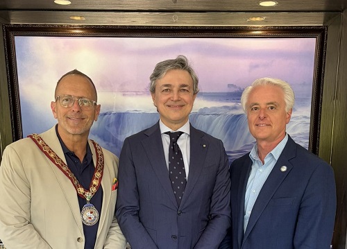 Three men in formal attire posing for a photo; left wears a ceremonial chain, middle in a dark suit, right in a blue blazer, with a waterfall painting behind.