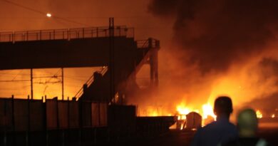 Night scene of a warehouse on fire with large flames and dark smoke; silhouettes of onlookers stand nearby as emergency lights glow in the flames.