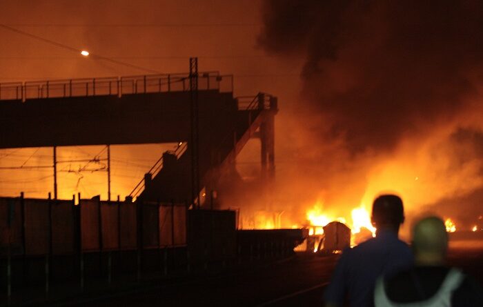Night scene of a warehouse on fire with large flames and dark smoke; silhouettes of onlookers stand nearby as emergency lights glow in the flames.