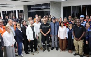Large diverse group of professionals standing together in a modern indoor facility, some in uniforms, posing for a group photo on camera.