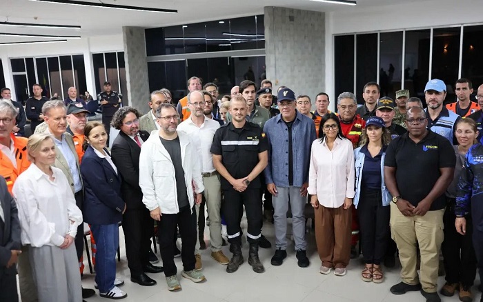 Large diverse group of professionals standing together in a modern indoor facility, some in uniforms, posing for a group photo on camera.
