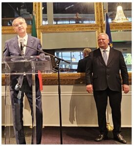 Two men in suits at a podium during a formal press conference; one speaks at the microphone, the other stands nearby in a room with gold-framed mirrors and a flag behind.