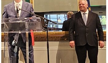 Two men in suits at a podium during a formal press conference; one speaks at the microphone, the other stands nearby in a room with gold-framed mirrors and a flag behind.