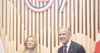 Two politicians shake hands in front of a wooden backdrop with a large G7 logo behind them, flags on either side.
