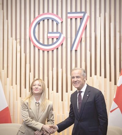 Two politicians shake hands in front of a wooden backdrop with a large G7 logo behind them, flags on either side.
