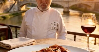 Chef seated at an outdoor riverside table with a plate of tomato pasta and a glass of red wine at sunset over a city bridge.