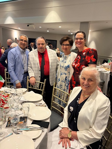 Group of six adults at a banquet table in a reception hall, posing for a photo; everyone is dressed in smart attire and the table is set with plates, glasses, and napkins.