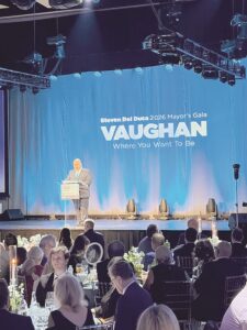 Politician delivering a speech at a formal gala on a stage with a blue backdrop reading 'VAUGHAN' and 'Where You Want To Be'.