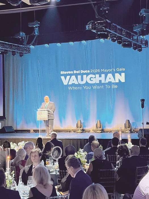 Man at a podium on a blue gala stage with a 'VAUGHAN' banner reading 'Steven Del Duca 2026 Mayor’s Gala' during a formal banquet.