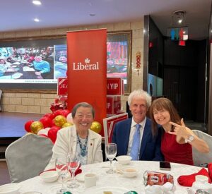 Three people pose at a banquet table under a red Liberal banner; balloons decorate the scene, with two women and a man in a suit smiling at the camera.