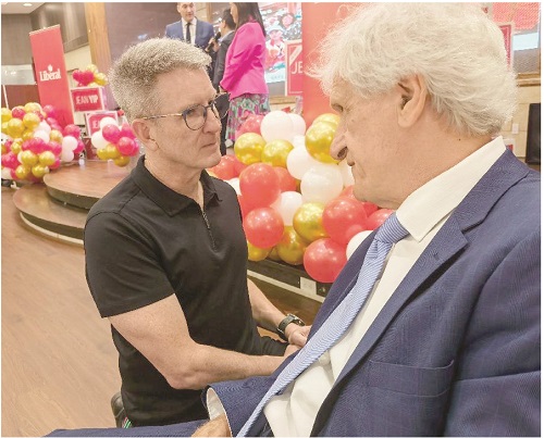 Two men talk closely at an event in a shop, with red, yellow, and white balloons in the background.