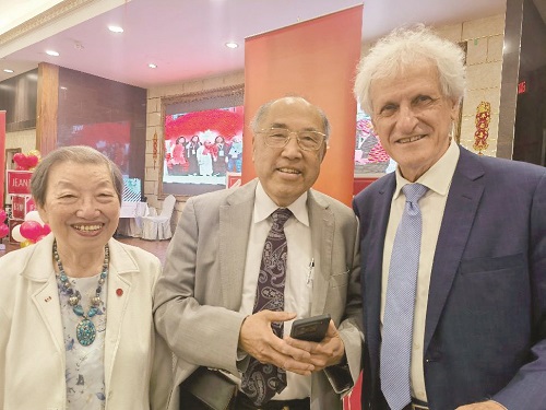 Three smiling adults posing for a photo at a formal event: a woman in a white jacket with a turquoise necklace, a man in a light suit holding a phone, and a man in a navy suit.