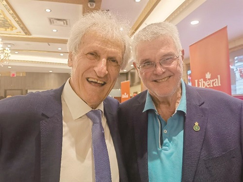 Two men in blazers smiling for a selfie at a Liberal party event, indoor venue with banners in the background.
