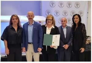 Five professionals posing on a stage, center woman holding a green certificate folder; backdrop shows repeating circular logos.