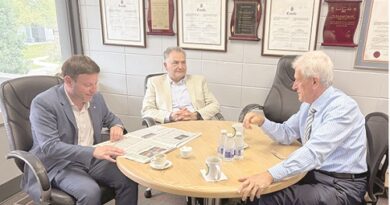 Three men in a business meeting around a round table with cups, in a room with framed certificates on the wall behind them.