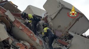 Three firefighters in yellow gear climb over rubble from a collapsed building, using ropes; Vigili del Fuoco logo in the corner.