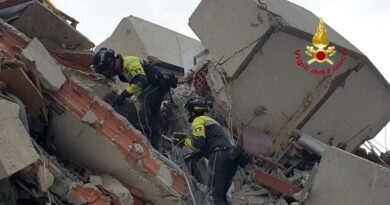 Three firefighters in yellow gear climb over rubble from a collapsed building, using ropes; Vigili del Fuoco logo in the corner.