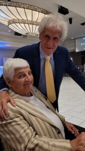 Older woman seated and smiling, wearing a beige striped jacket, with a man in a blue suit and yellow tie standing beside her and resting a hand on her shoulder in a bright lobby.
