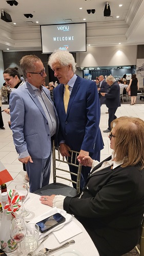 Two men in suits talk at a round dining table during a formal event; a woman sits nearby as a 'WELCOME' screen glows in the background.