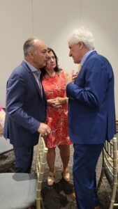 Three adults stand in a small circle indoors: two men in blue suits speaking to each other while a woman in a red floral dress watches, all engaged in a close, serious conversation.
