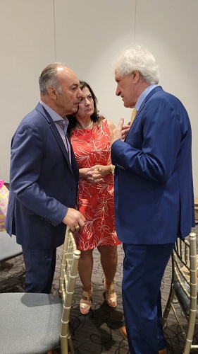 Three adults stand in a small circle indoors: two men in blue suits speaking to each other while a woman in a red floral dress watches, all engaged in a close, serious conversation.