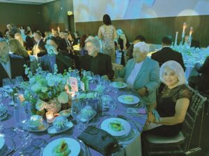 Elegant round-table dinner with six guests in formal attire, centerpiece flowers, candles, and plated meals at a gala event.