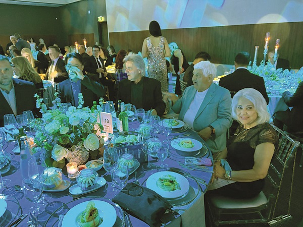 Elegant round-table dinner with six guests in formal attire, centerpiece flowers, candles, and plated meals at a gala event.