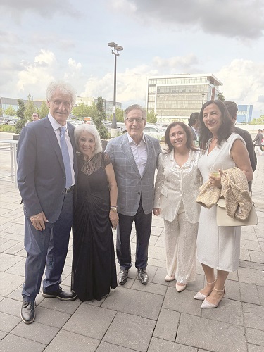 Six adults in formal attire pose for a group photo outdoors on a paved plaza, smiling at the camera.