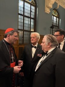 Cardinal in red hat conversing with three men in tuxedos at a formal event in a room with tall arched windows.