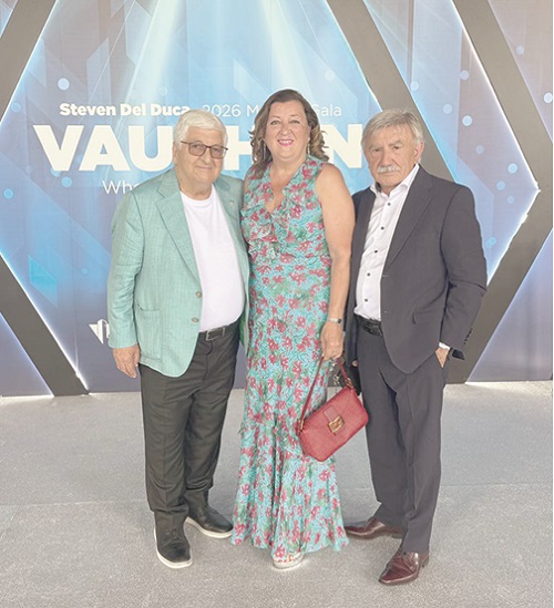 Three adults posing for a photo at an event: a man in a light green blazer, a woman in a floral dress with a red purse, and a man in a dark suit, in front of a blue geometric backdrop.