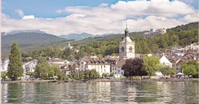 Lakeside town with a white church and clock tower along a calm lake, hills and mountains in the distance.
