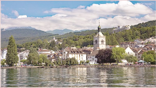 Lakeside town with a white church and clock tower along a calm lake, hills and mountains in the distance.