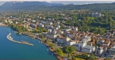 Aerial view of a lakeside town with buildings along the shore, a marina, and green hills in the background.