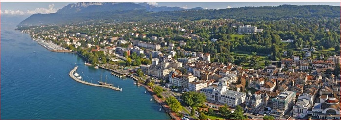 Aerial view of a lakeside town with buildings along the shore, a marina, and green hills in the background.