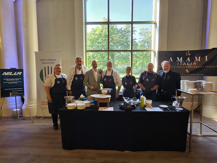 Group of eight chefs in white shirts and dark aprons stand behind a black kitchen prep table with bowls and utensils in a bright event room; banners on either side hint at a culinary demo.