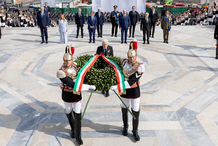Two guards in ornate uniforms present a wreath with Italian flag ribbons at a formal state ceremony, with officials in suits behind them.