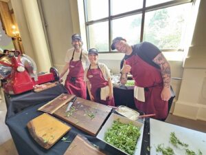 Three people in red aprons at a kitchen demo table, chopping herbs beside a red meat slicer on display.
