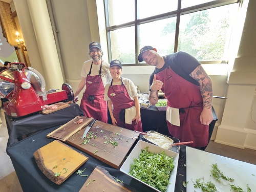 Three people in red aprons at a kitchen demo table, chopping herbs beside a red meat slicer on display.