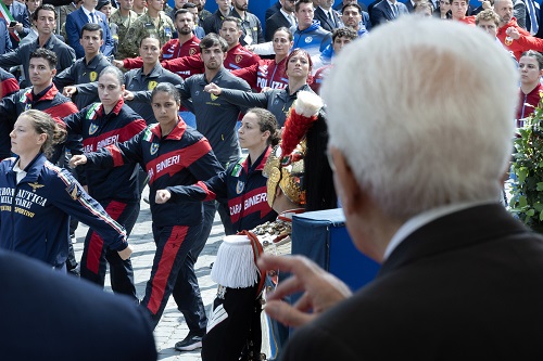 Back of an elderly man in a suit addressing a crowd of uniformed marchers at a ceremony around him far in the background.
