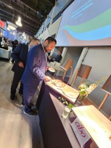 Man in a blue suit samples food at a conference buffet, with a projector screen and attendees in the background.