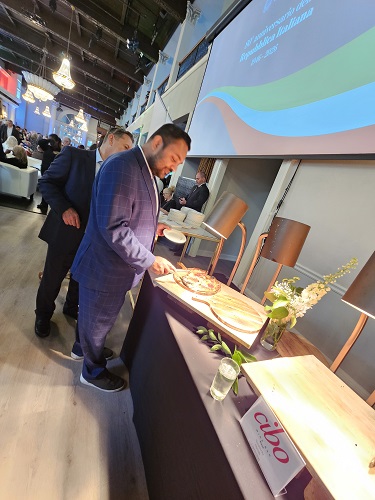 Man in a blue suit samples food at a conference buffet, with a projector screen and attendees in the background.