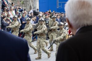 Soldiers in camouflage march in formation during a city parade while a crowd of onlookers watches from the sides.