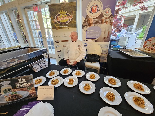 Chef in a white coat seated behind a table with many plates of pasta samples at a food expo.