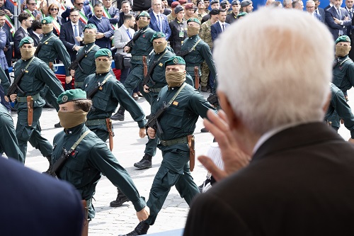 Soldiers in green uniforms march in formation with blindfolds during a parade, as spectators watch from the stands and street.